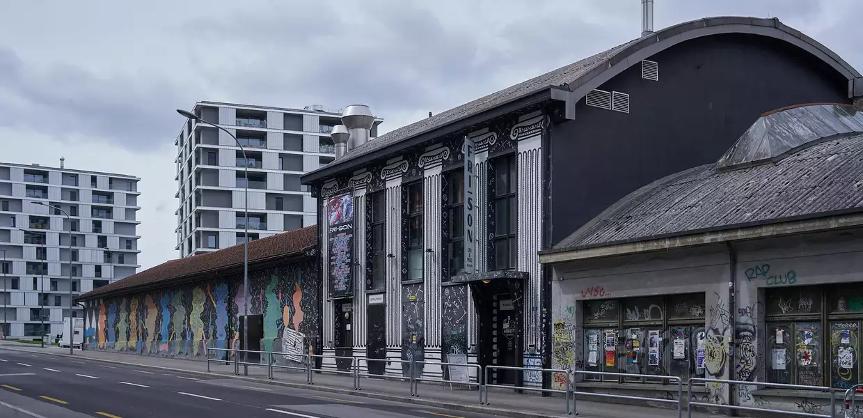 Vue du bâtiment et de l'entrée depuis le côté opposé de la rue. Deux immeubles se trouvent à l'arrière-plan. 